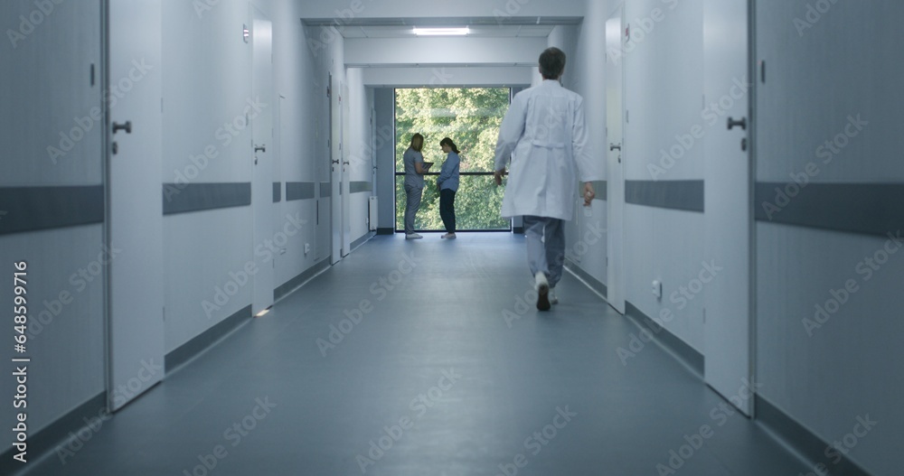 Clinic corridor: Doctors and professional medics walk. Nurse with digital tablet comes to elderly woman standing near window. Medical staff and patients in modern hospital or medical center hallway.