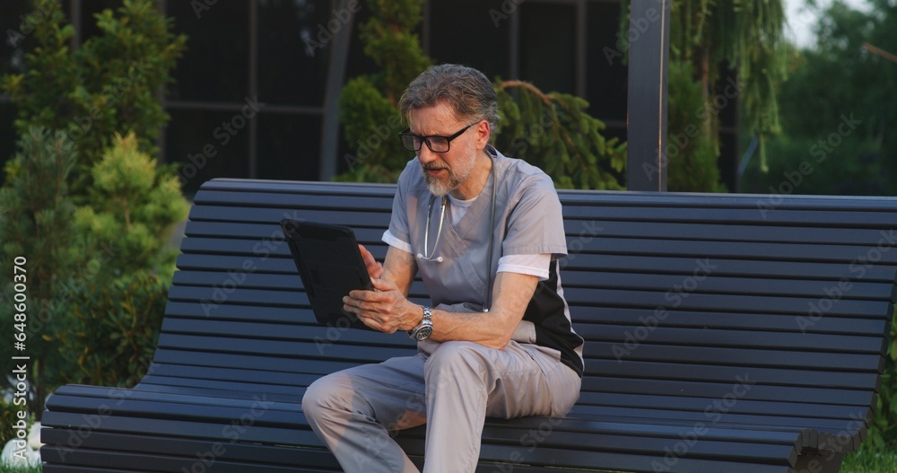 Fototapeta premium Doctor in uniform sits on bench outside modern hospital, talks to patient about treatment during online appointment using digital tablet. Medical staff of clinic or medical center. Healthcare system.
