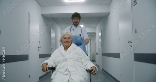 Young doctor in glasses carries senior patient in wheelchair down hospital corridor. Old woman talks to medic and smiles. Mature doctor asks female patient about her health. Medical staff at work.