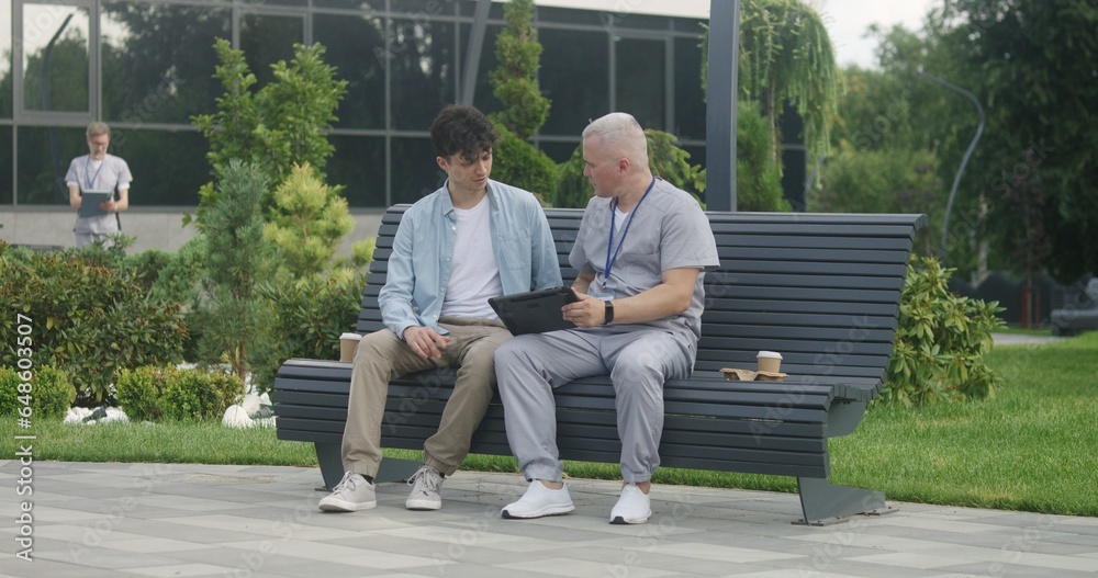 Fototapeta premium Doctor with tablet in hands sits with adult patient on bench and suggests treatment to him. Professional in medical uniform makes diagnosis to client using digital tablet. Medical staff work outdoor.