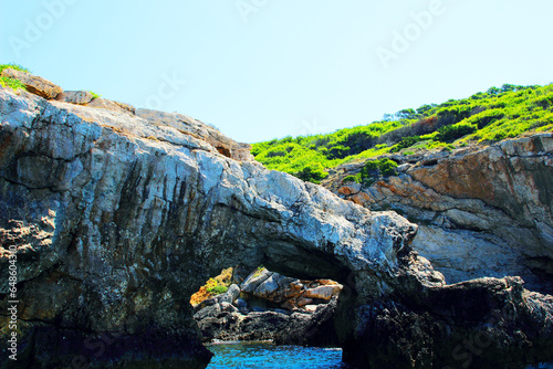 Multi-level scenery from Tremiti Islands (Isole Tremiti) with a cove of iridescent blue Adriatic Sea rippling under a rocky tunnel, opening to momentous ridges of green vegetation and fierce stone