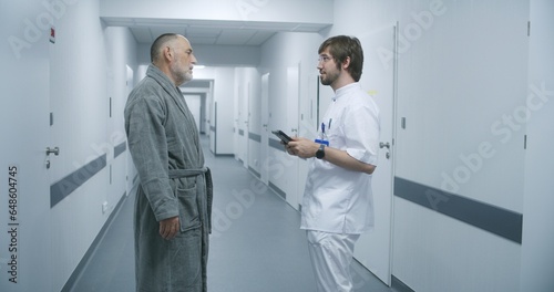 Tracking shot of doctor with digital tablet walking along clinic corridor with elderly man. Healthcare specialist and patient talk  discuss treatment. Medical staff and patients in hospital hallway.