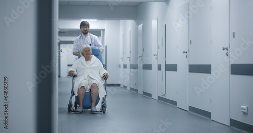 Male nurse pushes wheelchair with female patient in clinic corridor. Female doctor comes to colleague and elderly patient, talks, uses digital tablet. Medical staff and patients in hospital hallway.