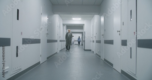 Multiracial medical staff in uniform walk clinic corridor. Elderly patient goes to appointment with doctor. Health care specialists greet in medical center hallway. Dolly shot. Back view. Slow motion.