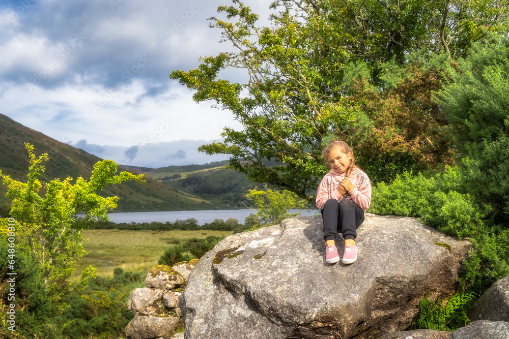 Young girl sitting on a large rock and smiling to camera with lake Lough Dan and mountain valley in background, Wicklow Mountains, Ireland
