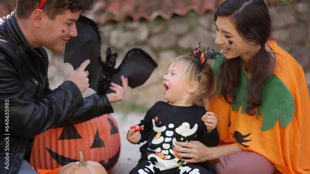 Multinational family with a child dressed up for Halloween in costumes and weighed play together the background of Halloween props, bats, pumpkins, skulls, spiders, brooms, burning candles, cobwebs.