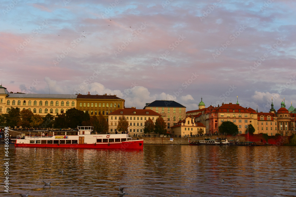 Cityscape with historic old buildings with spires and red roofs, and trees on the riverbank with red boat. Autumn, late evening, sunset. Prague, Czech Republic, Vltava River, October 2022