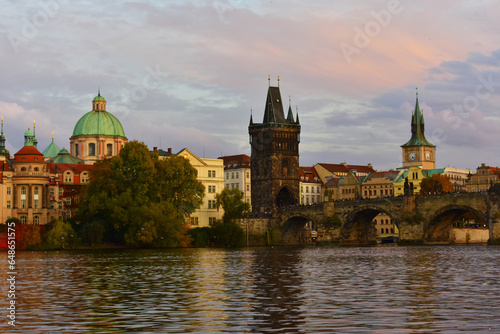 Cityscape with historic old buildings with green dome and red roofs, trees on the riverbank. Charles Bridge over the Vltava River. Autumn, late evening, sunset. Prague, Czech Republic, October 2022
