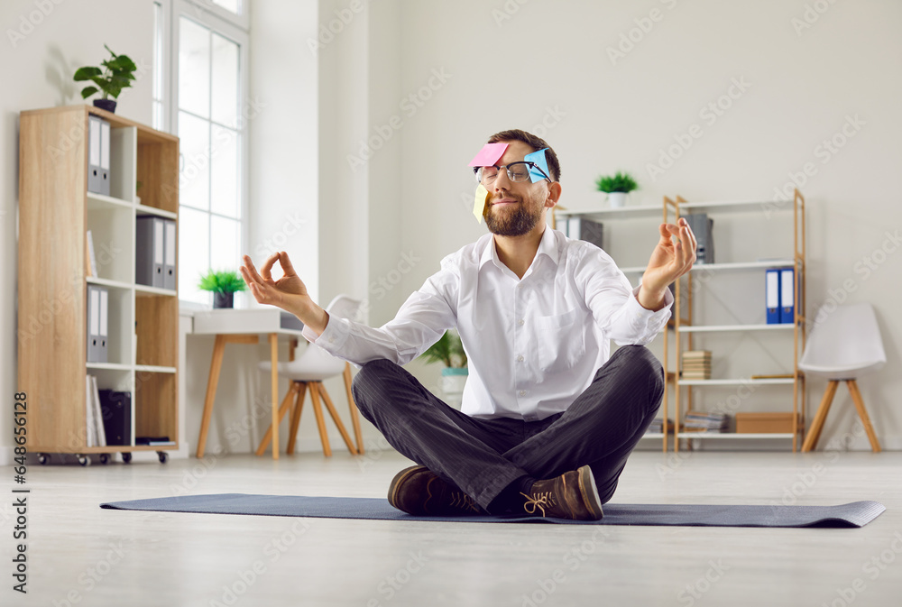 Fototapeta premium Funny busy man with sticky reminder notes on face sitting on yoga mat floor in his business office, doing meditation, and relaxing. Stress management, relaxation at work concept