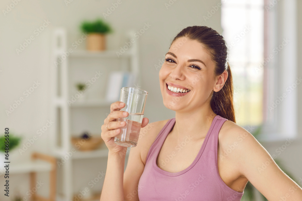 Portrait of a young sporty happy woman standing at home after working out with a glass of water, looking cheerful at the camera and smiling. Sport, healthy nutrition and lifestyle concept. Portrait of a young sporty happy woman standing at home after working out with a glass of water, looking cheerful at the camera and smiling. Sport, healthy nutrition and lifestyle concept.