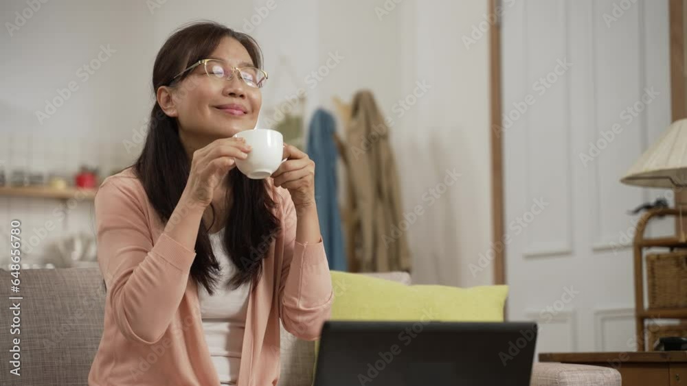 happy asian middle aged businesswoman enjoying aroma while taking coffee break from work on the computer in the living room at home happy asian middle aged businesswoman enjoying aroma while taking coffee break from work on the computer in the living room at home