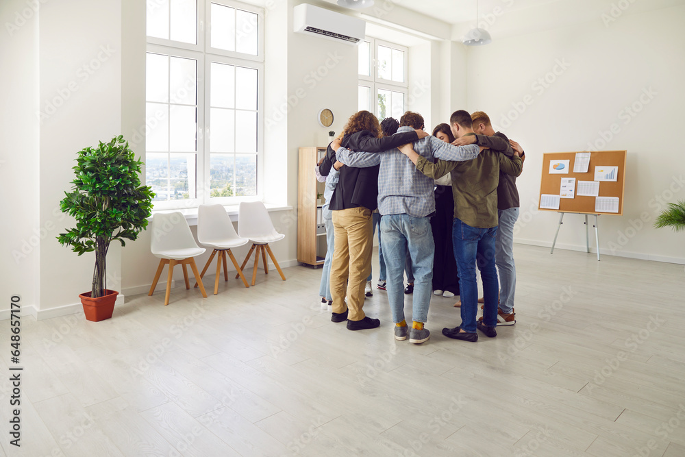 Group of a young business people company employees men and women in casual clothes standing in a circle and hugging indoors standing in office in a meeting room. Team work concept. Group of a young business people company employees men and women in casual clothes standing in a circle and hugging indoors standing in office in a meeting room. Team work concept.