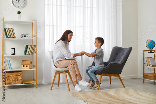 Cute boy and female psychologist holding hands together. Little patient and doctor sitting in front of each other during psychological session. Medical consultation, psychological support