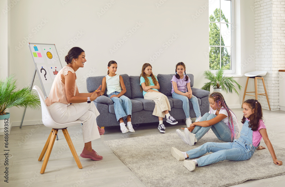 Smiling school psychologist specialist together with group of little kid children friend girls, sitting in office classroom after class, discuss bad behaviour and find solution for conflict situation Smiling school psychologist specialist together with group of little kid children friend girls, sitting in office classroom after class, discuss bad behaviour and find solution for conflict situation