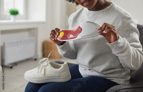 Bild auf Leinwand Cropped shot of young African American woman with shoe on her lap sitting on chair at home and looking at orthotic arch support insole that she is holding in her hands