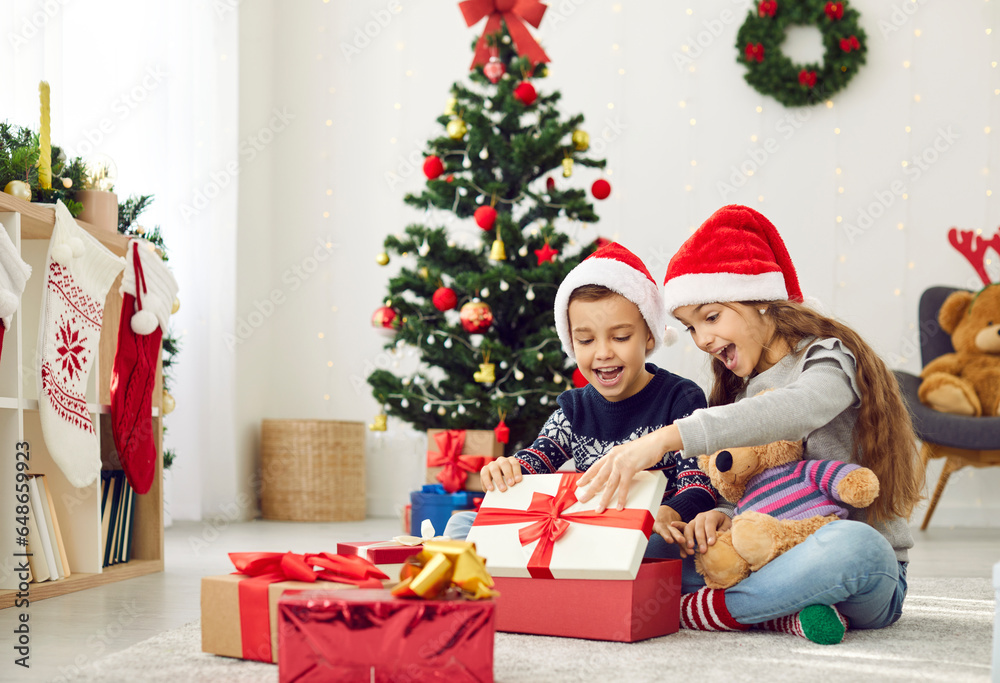 Happy little children opening presents on Christmas morning. Two excited kids in red hats sitting on floor in beautiful, decorated living room and together looking inside box with wonderful Xmas gift Happy little children opening presents on Christmas morning. Two excited kids in red hats sitting on floor in beautiful, decorated living room and together looking inside box with wonderful Xmas gift