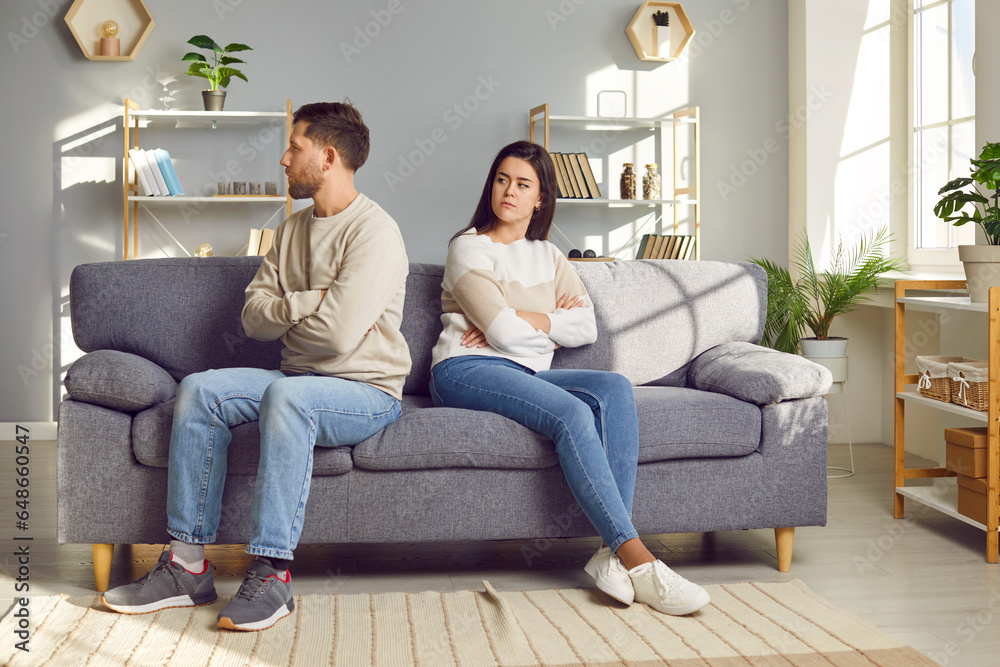 Family couple having conflict. Young man and woman quarrel and don't talk to each other. Two angry people sit down on sofa, turn away from each other, and keep sitting back to back, ignoring one other