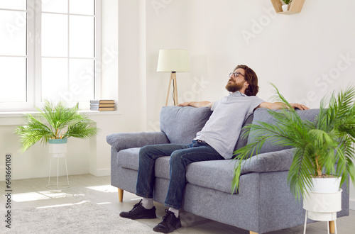 Man is relaxing on the couch at home. Bearded man in polo shirt, jeans and glasses is enjoying a quiet day off and sitting on a comfortable grey sofa in a living room with white walls and green plants
