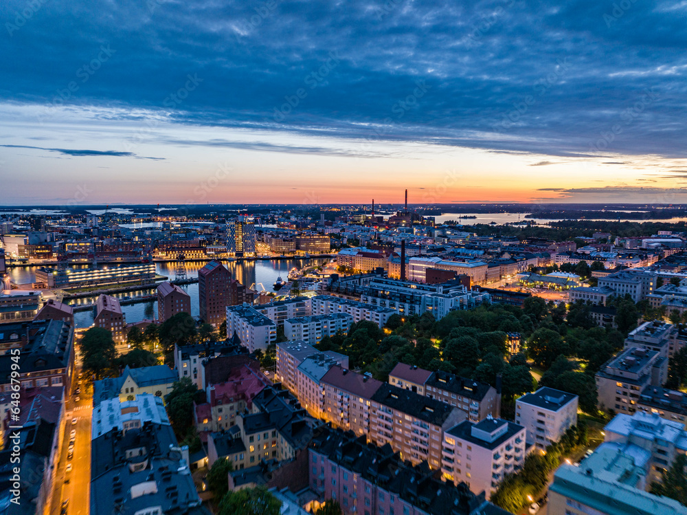 Aerial shot of illuminated buildings in residential urban borough and shipyard in bay. Evening cityscape against romantic colour twilight sky. Helsinki, Finland