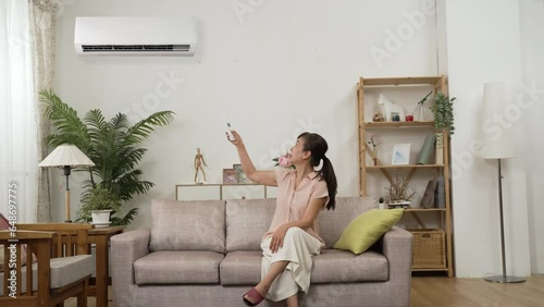 sweating asian mature woman feeling dehydration during menopause and fanning with hands. she switches on air conditioner and enjoys cool air with spreading arms on sofa at home