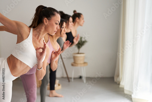 Group of Caucasian young people practicing yoga asanas indoors. Concentrated sports women performing exercises in class. Fitness girls stretching. Healthy lifestyle in community and daily routines.  photo