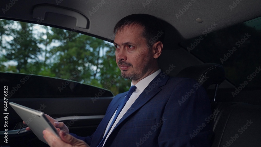 Serious businessman uses computer tablet to obtain business information in interior of business car. Business man works in car. Politician works and looks at information on tablet in car. Man in suit