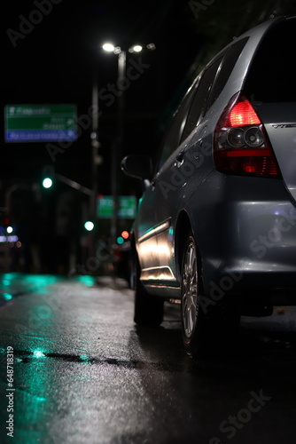 Carro com luzes acesas, estacionado na calçada em uma rua refletida em uma noite chuvosa