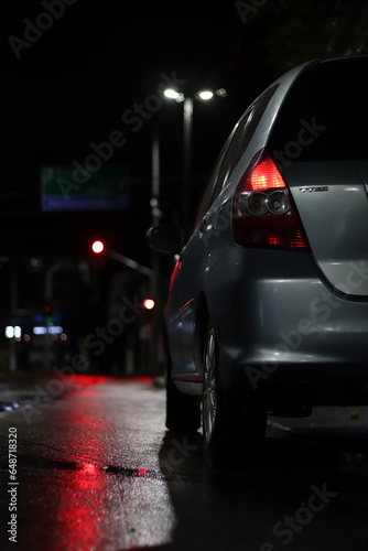 Carro com luzes acesas, estacionado na calçada em uma rua refletida em uma noite chuvosa