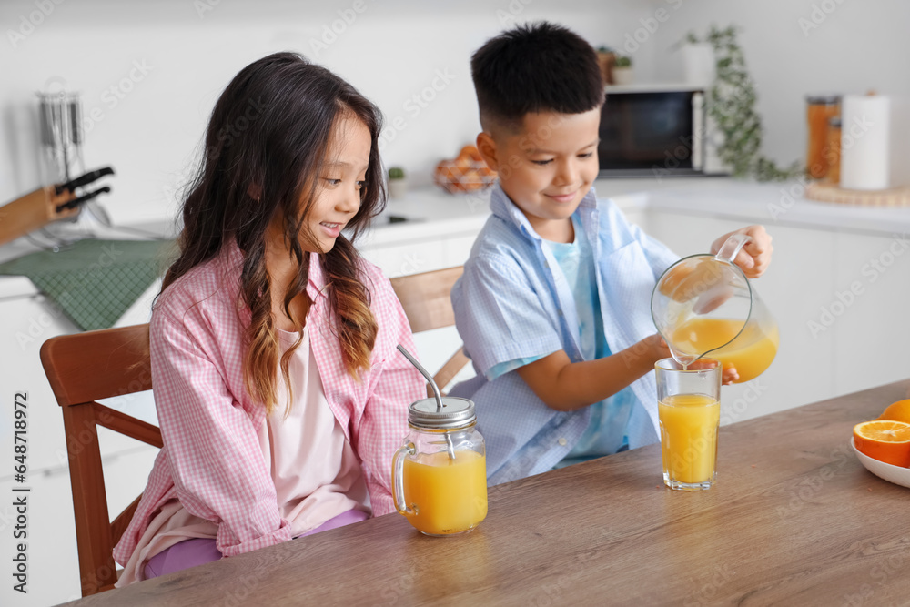 Little Asian children pouring fresh citrus juice into glasses from jug at table in kitchen Little Asian children pouring fresh citrus juice into glasses from jug at table in kitchen