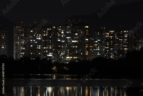 Janelas de vários prédios acesas durante a noite, sendo refletidas em um lago photo