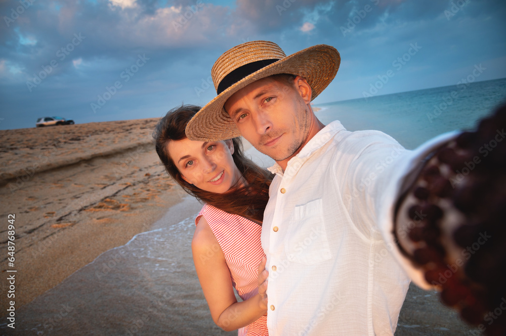 Close-up of smiling happy young couple, two family man and woman hugging each other, taking first person selfie at sunrise or sunset on sea beach on summer day