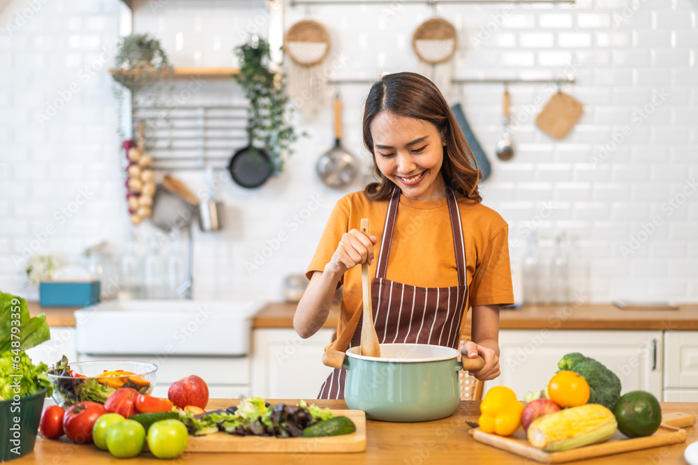 Young woman standing near stove and cooking, housewife, meal, chef, food.Happy woman looking and smelling tasting fresh delicious from soup in a pot with steam at white interior kitchen Young woman standing near stove and cooking, housewife, meal, chef, food.Happy woman looking and smelling tasting fresh delicious from soup in a pot with steam at white interior kitchen