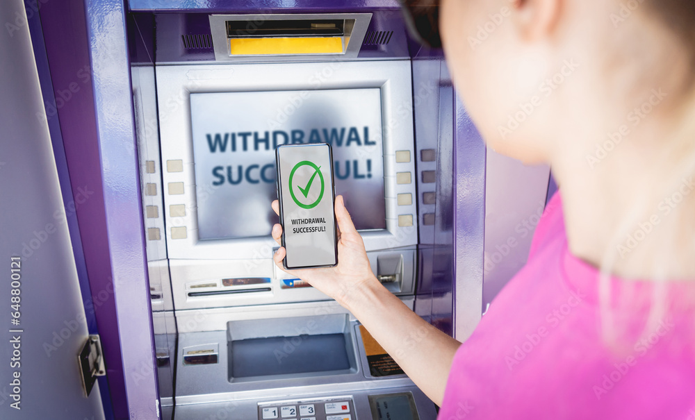 Young woman using the smart phone for cardless withdrawing the cash near the ATM Young woman using the smart phone for cardless withdrawing the cash near the ATM