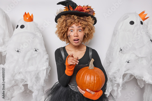 Indoor photo of young surprised African american female in costume of witch holding pumpkin standing in centre surrounded by spooky ghosts seeing something unusual or unexpected pointing at you photo