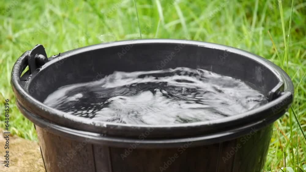 The rain falls into the bucket and creates splashes, Rain drops into the water bucket, black plastic bucket filled water from trickle leaking water spilled on the floor, liquid container The rain falls into the bucket and creates splashes, Rain drops into the water bucket, black plastic bucket filled water from trickle leaking water spilled on the floor, liquid container