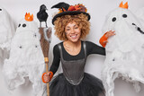 Holiday concept. Indoor waist up of young cheerful smiling broadly African american lady wearing costume of witch holding broom standing in centre on white background surrounded by spooky ghosts