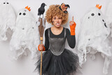 Halloween celebration. Studio shot of young angry African american lady wearing costume of witch holding broom with raven standing in centre on background of ghosts pointing to top having party