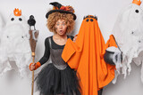 Shocked curly haired young woman poses near orange ghost holds broom with crow prepares for Halloween holidays surrounded by spooky creatures isolated over white background being in festive mood