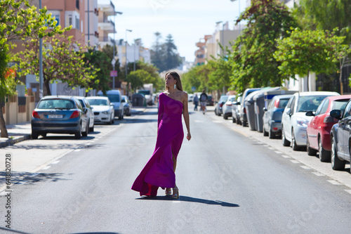 Fotografi Young beautiful blonde woman dressed in purple dress walks along a large lonely avenue among the stopped cars