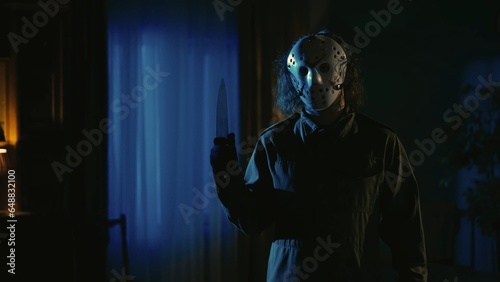 Photos Man maniac in white hockey mask standing in the dark living room holding knife in hand, looking at the camera
