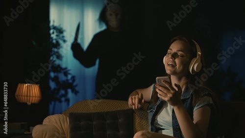 Tableau sur toile Girl in headphones sitting on the sofa in the living room, listening to music, man maniac in white mask standing with knife near window behind