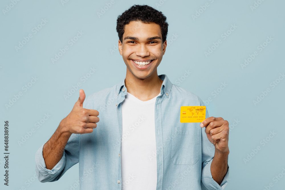 Young smiling man of African American ethnicity he wear shirt casual clothes hold in hand mock up of credit bank card show thumb up isolated on plain pastel light blue cyan background studio portrait. Young smiling man of African American ethnicity he wear shirt casual clothes hold in hand mock up of credit bank card show thumb up isolated on plain pastel light blue cyan background studio portrait.