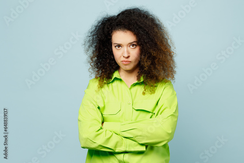 Young frowning grumpy sad latin woman she wear green shirt casual clothes hold hands crossed folded look camera isolated on plain pastel light blue cyan background studio portrait. Lifestyle concept.