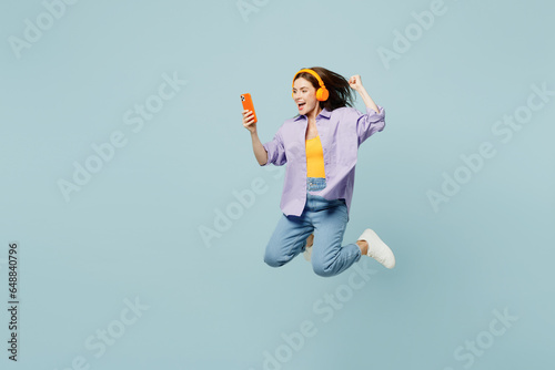 Full body side view young happy woman she wears purple shirt yellow t-shirt casual clothes jump high use mobile cell phone listen to music in headphones isolated on plain pastel light blue background.