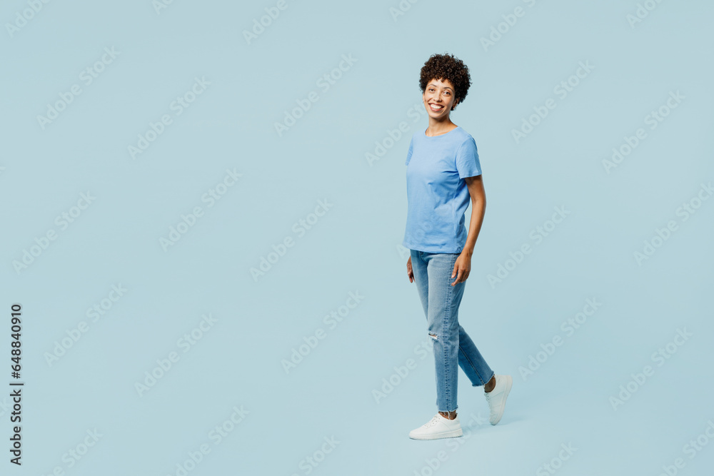 Full body side profile view young fun woman of African American ethnicity wear t-shirt casual clothes walking going look camera isolated on plain pastel light blue cyan background. Lifestyle concept. Full body side profile view young fun woman of African American ethnicity wear t-shirt casual clothes walking going look camera isolated on plain pastel light blue cyan background. Lifestyle concept.