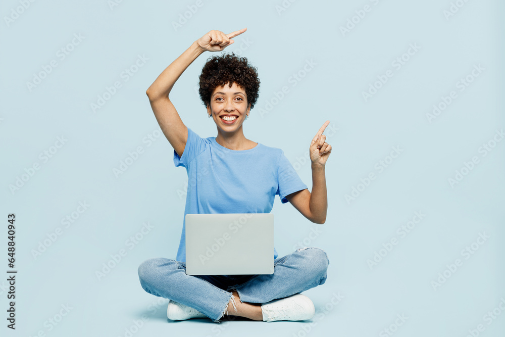 Full body young IT woman of African American ethnicity wear t-shirt casual clothes sit hold use work on laptop pc computer point aside isolated on plain pastel blue cyan background Lifestyle concept. Full body young IT woman of African American ethnicity wear t-shirt casual clothes sit hold use work on laptop pc computer point aside isolated on plain pastel blue cyan background Lifestyle concept.