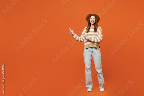 Full body young woman she wearing striped sweater hat casual clothes point index finger aside indicate on workspace area copy space mock up isolated on plain orange red color wall background studio.