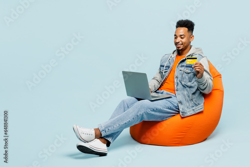 Full body young IT man of African American ethnicity wear denim jacket orange t-shirt sit in bag chair use laptop pc computer credit bank card shopping online isolated on plain blue background studio