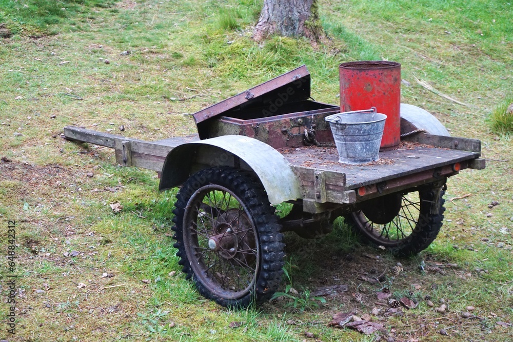 Fototapeta premium Vintage homebuilt trailer with spoked knobby tread wheels hauling a galvanized bucket, square metal box with open lid and tall red painted tin container