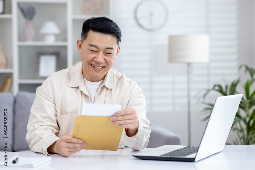 Happy and successful asian received mail notification envelope with good news, man sitting at table at home, working in home office in living room, joyfully celebrating winning news. Happy and successful asian received mail notification envelope with good news, man sitting at table at home, working in home office in living room, joyfully celebrating winning news.